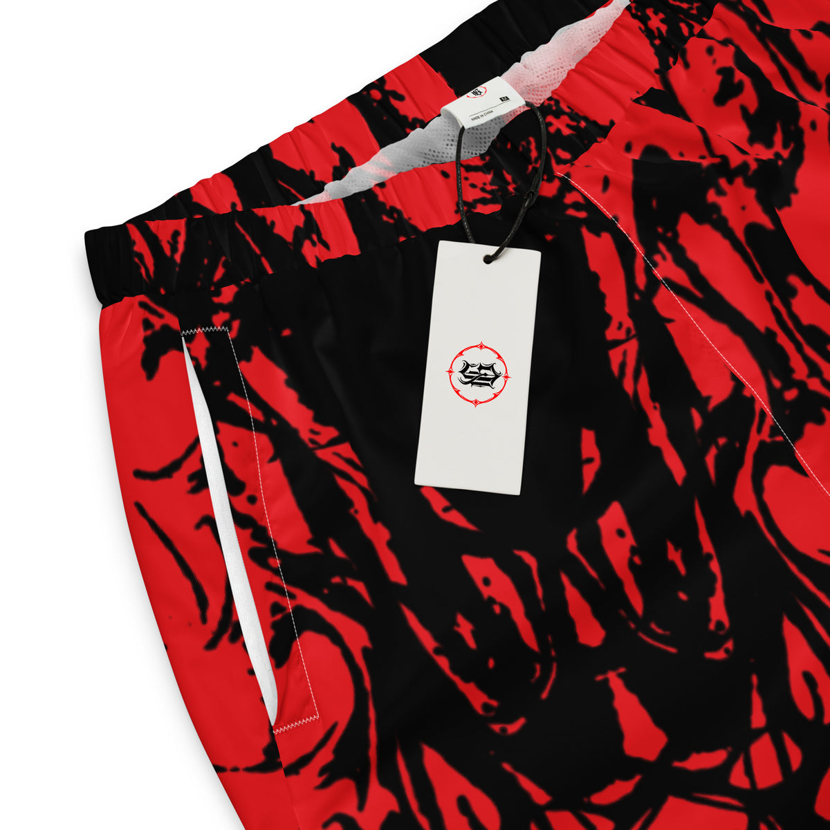 Close-up of red and black patterned pants with a white hangtag featuring a circular logo, photographed on a white background.