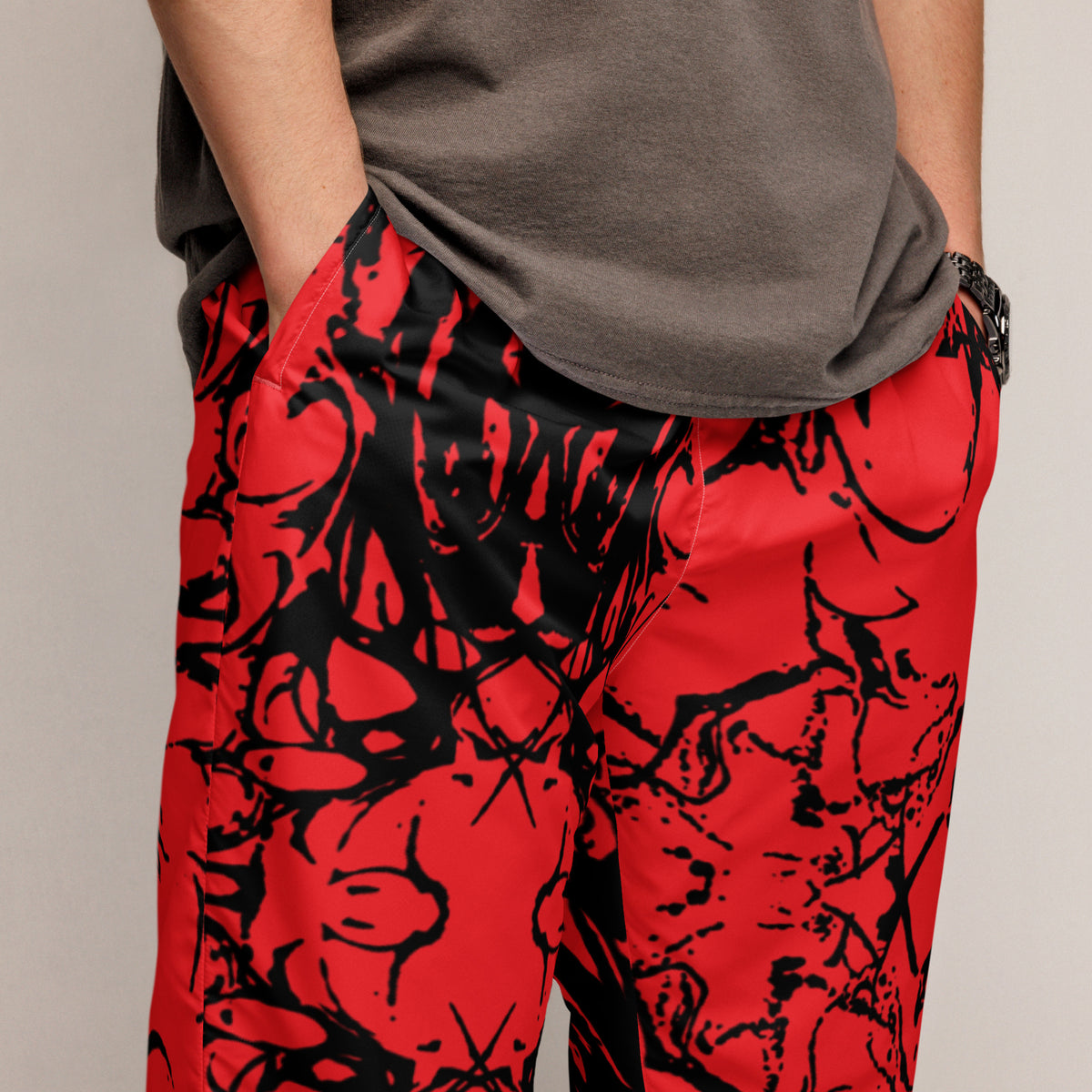 A close-up of a person wearing bright red pants with a bold black abstract graffiti-style print, paired with a taupe-gray t-shirt, against a neutral light gray background.