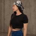 Woman wearing Ink-Riot Bandana as a headband, black and white all-over print, styled with a black crop top and jeans against a concrete wall