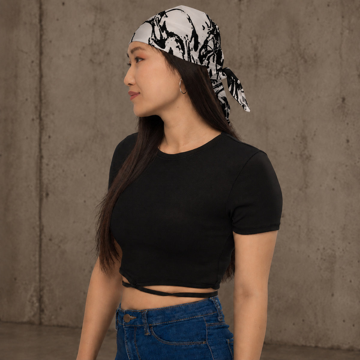 Woman wearing Ink-Riot Bandana as a headband, black and white all-over print, styled with a black crop top and jeans against a concrete wall