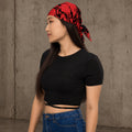 A woman wearing a red and black patterned bandana headscarf, a black short-sleeve crop top, and blue jeans, standing against a textured concrete wall.