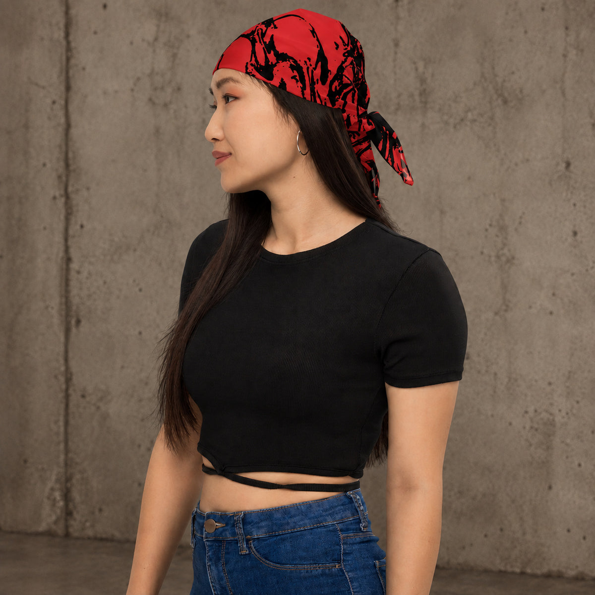 A woman wearing a red and black patterned bandana headscarf, a black short-sleeve crop top, and blue jeans, standing against a textured concrete wall.