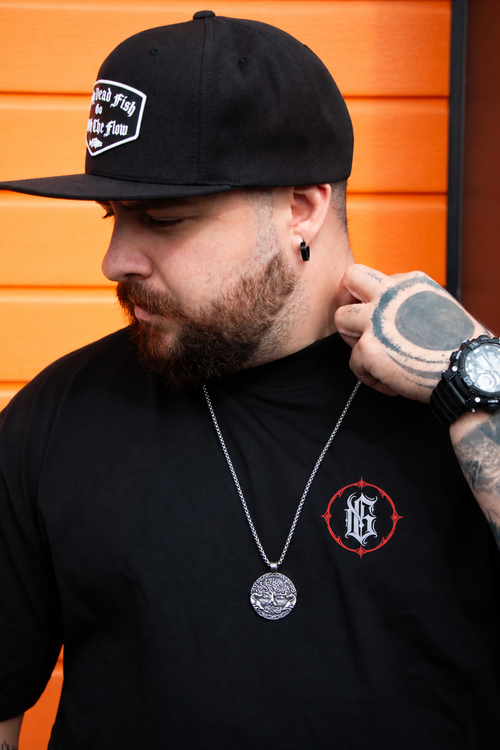 A man wearing a black baseball cap with a white patch logo, a black t-shirt featuring a red and white circular 'G' emblem on the left chest, and a silver pendant necklace, posed against an orange-paneled wall.